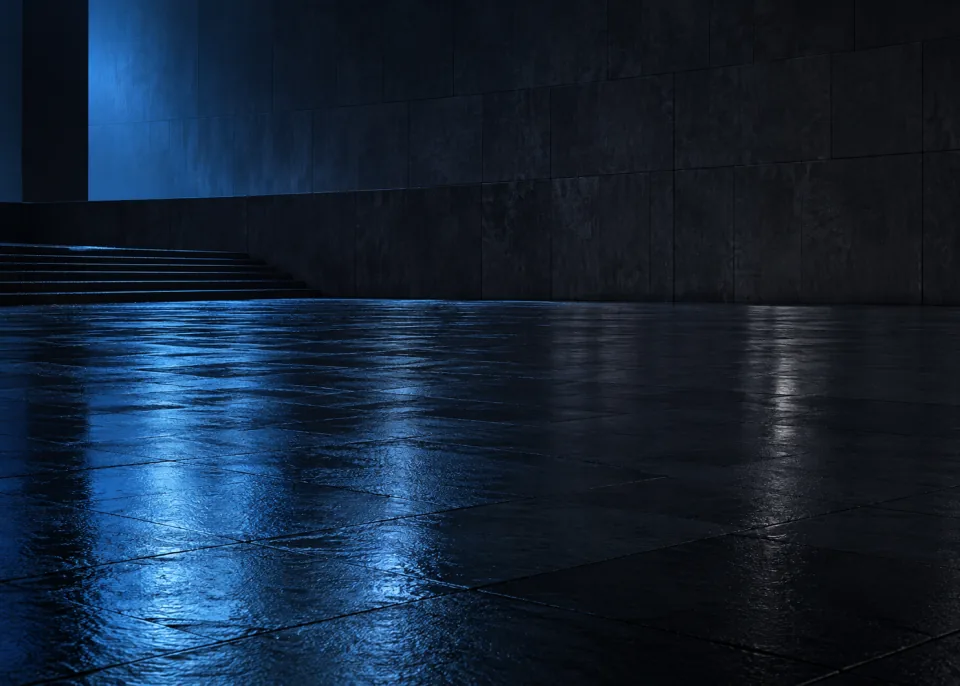 Rain on Slate: Low view of an empty rain-wet dark slate plaza reflecting one broad muted blue light field from outside frame, simple shadowed wall background.