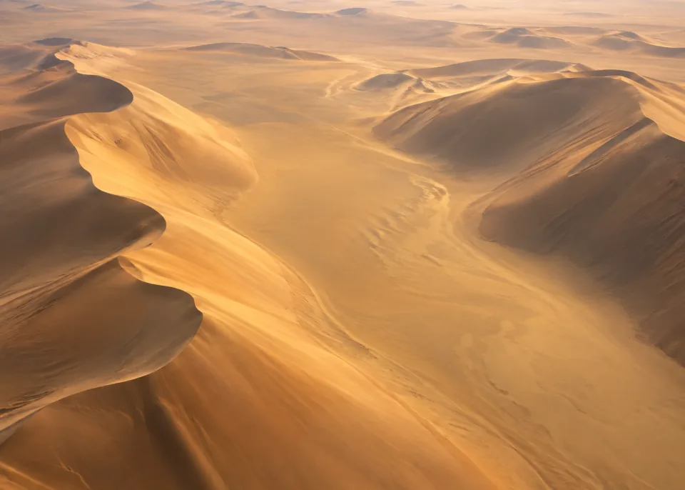 Amber Valley: High oblique aerial of an enormous golden sand trough flanked by smooth curved dunes, soft diffuse light, broad textureless background.