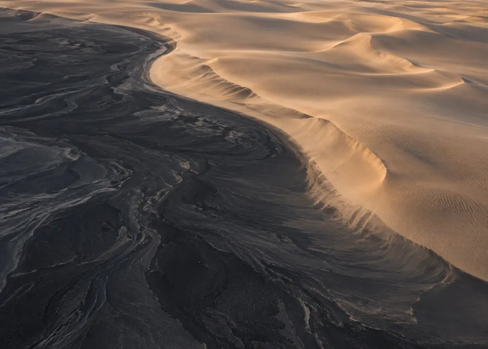 Black Sand: Minimal aerial meeting of dark volcanic sand and warm taupe windblown dunes in one broad curved boundary, soft late light.