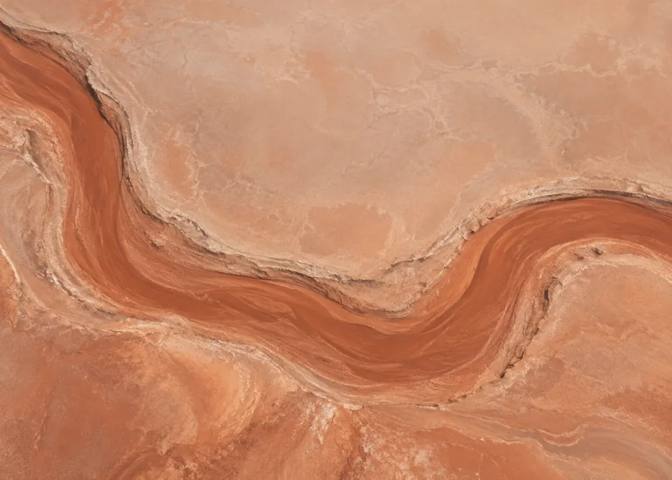 Clay Riverbed: Top-down view of one broad dry terracotta river channel curving through a flat muted peach clay plain, subtle raised banks without branching clutter.