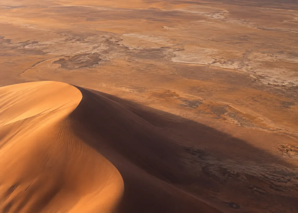 Desert Shadow: A monumental rounded sand dune cropped at bottom left casts a single long soft plum-brown shadow across muted amber terrain, low-detail upper field.