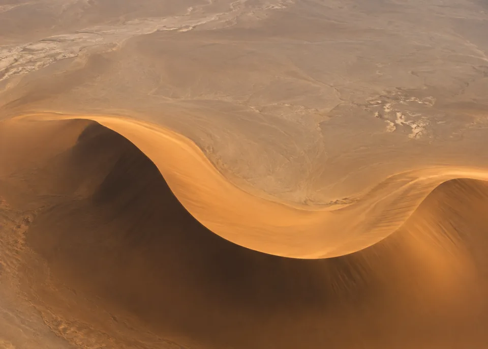 Dune S: Top-down aerial of one enormous ochre dune making a broad S-curve across lower half, soft umber shadow and uninterrupted muted sand expanse.