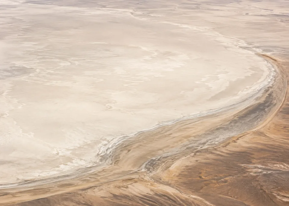 Ivory Basin: Vast warm ivory dry lakebed with a single broad muted beige shoreline arc crossing lower right, soft clay transitions, no cracked-earth grid.