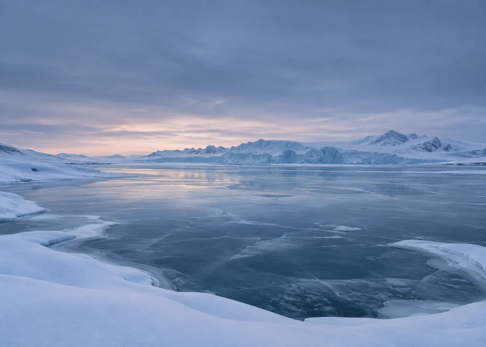 Frozen Lagoon: Low smooth snow-covered banks around an expansive slate-blue frozen lagoon, distant ice ridge, softly graded cold twilight sky.