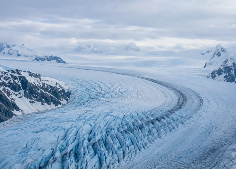 Glacier Curve: Oblique close aerial of one enormous sweeping glacier bend, broad smooth pale blue ice regions and a few dark blue creases concentrated in lower half.
