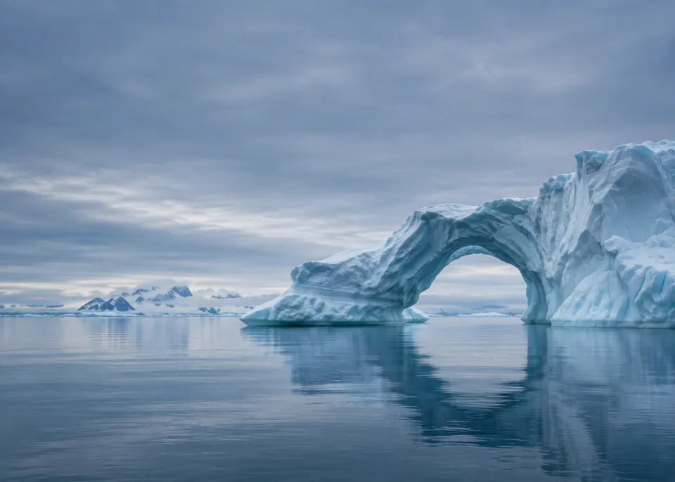 Ice Arch: One large low sculptural natural ice arch near lower right on still sea, pale muted cyan light, broad quiet slate-blue sky and water.