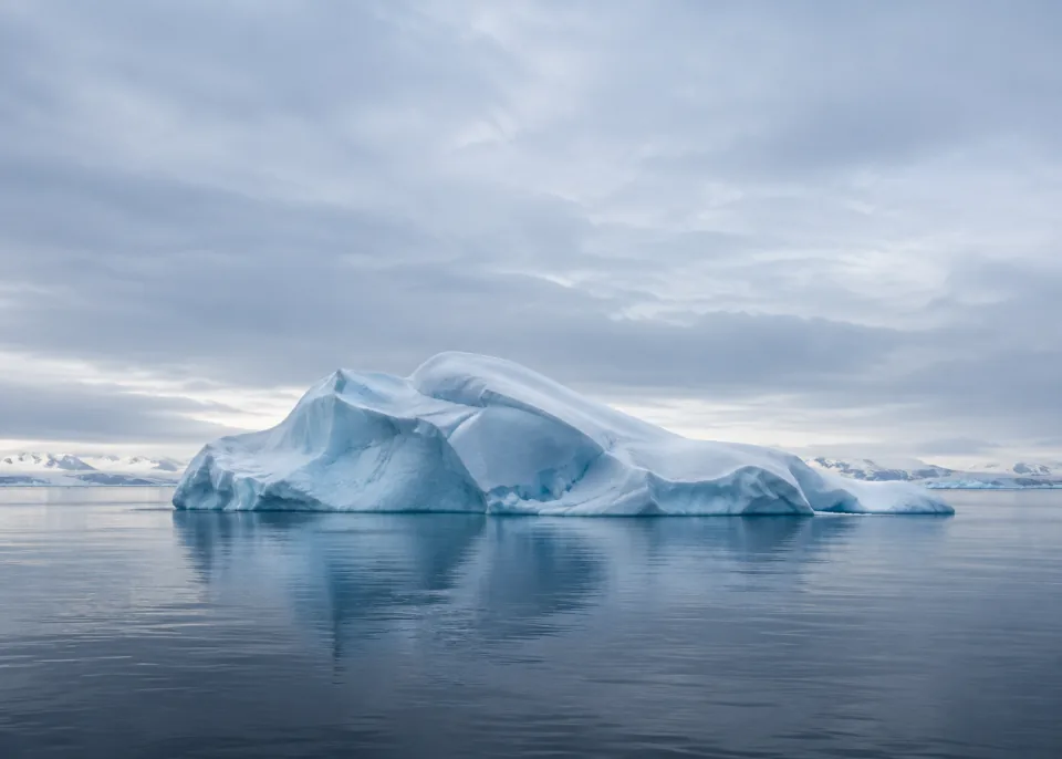 Iceberg Drift: Single broad rounded blue-white iceberg drifting in still dark slate water below center, thin distant horizon and vast pale blue-gray overcast sky.