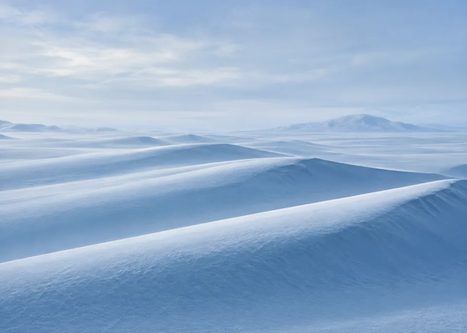 Snow Ridges: Minimal gentle snow dunes sculpted into broad smooth parallel ridges, cool blue shadows at lower edges, quiet powder-blue atmospheric upper field.