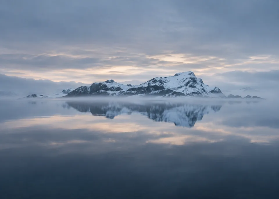 Winter Reflection: Distant low snow-covered rock island mirrored in glassy dark blue lake, thin fog across horizon, serene gray-blue dawn sky.