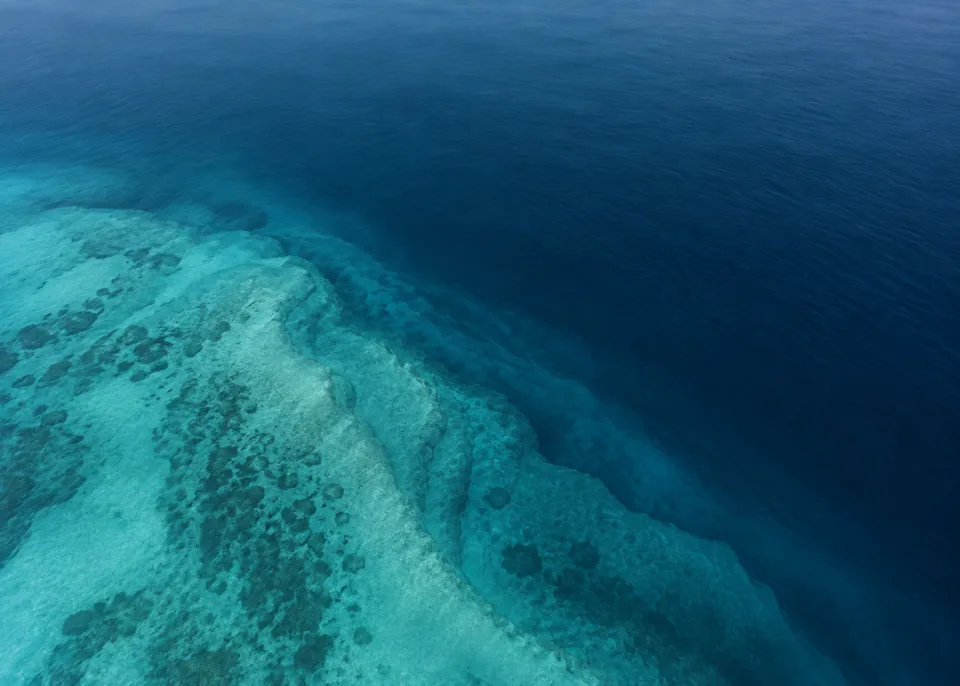 Atlantic Blue: Mostly deep slate blue ocean, one broad pale underwater ridge emerging softly from lower left and disappearing into navy depths, serene abstract aerial.