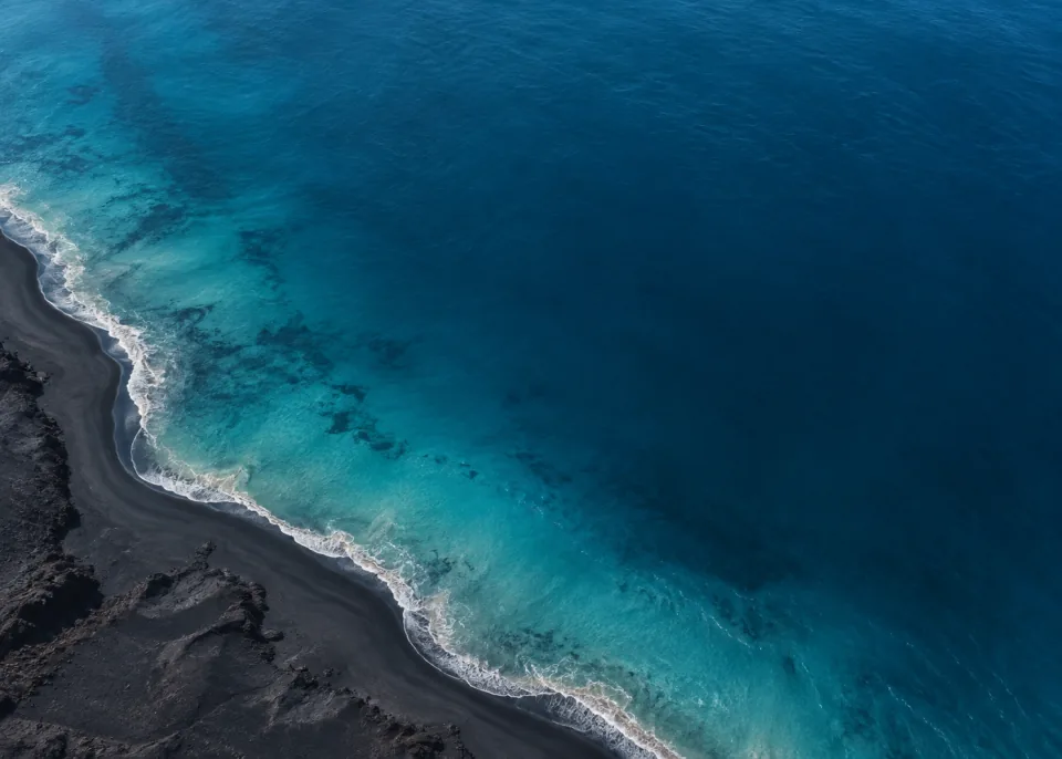 Black Shore: A single elegant curved black volcanic shoreline along lower left with soft subdued pale surf edge, deep muted blue ocean occupying most of frame.