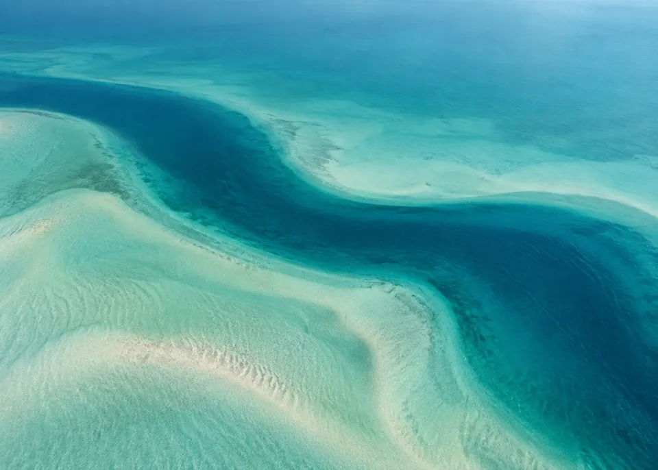 Lagoon Ribbon: One broad winding dark teal ribbon of deeper water crossing an expansive muted aquamarine lagoon, gently blurred sandy edges.