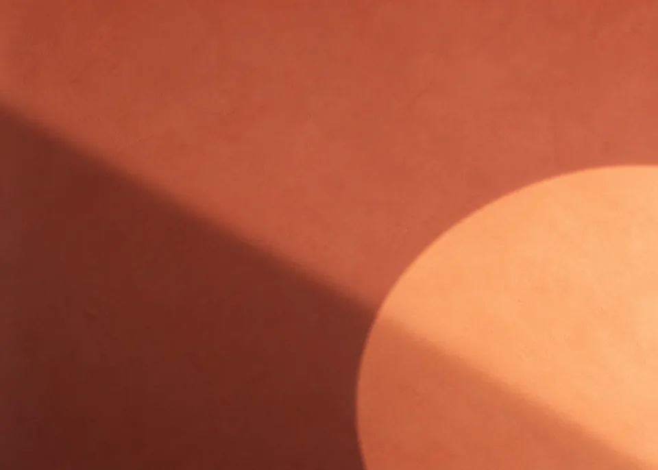 Circle of Dawn: A large soft-edged circular pool of muted apricot light falls partly out of frame near lower right on a dusty coral plaster wall, broad diagonal shadow.